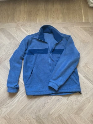 Blå fleecejacka från Columbia - Mysig blå fleecejacka från Columbia med dragkedja framtill och hög krage. Jackan har två sidofickor med dragkedja och en mörkare blå detalj över bröstet. Perfekt för kyliga dagar och enkel att matcha med andra plagg.