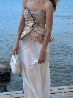 Champagnefärgad plisserad tubtopp - Säljer en unik champagnefärgad tubtopp med plisserade volanger och snygg struktur. Toppen är axelbandslös och har en lyxig känsla med metallicglans. Perfekt för dig som vill sticka ut med en statement piece. Endast använt en gång 