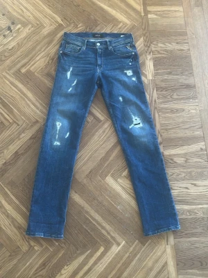 Blå slitna jeans från REPLAY ANBASS - Säljer ett par blå jeans från Replay  med snygga slitningar och distressed detaljer på både fram- och baksida. Klassisk femficksmodell med raka ben och normal midja. Slim fit passar upp til 170 cm
