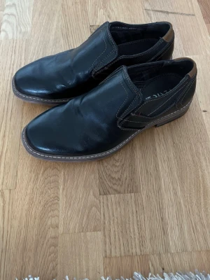Svarta loafers i skinn str. 42 - Säljer ett par svarta loafers  med bruna detaljer vid hälen och kontrastsömmar. Skorna har elastiska paneler på sidorna för enkel påtagning och en robust sula i brunt. Perfekta för en stilren look.