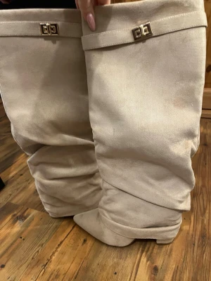 Beige slouchy stövlar med spänne - Snygga beige stövlar i mockaimitation med bred, slouchy passform och dekorativt guldfärgat spänne upptill. Klacken är ca 9cm, aldrig använda. 