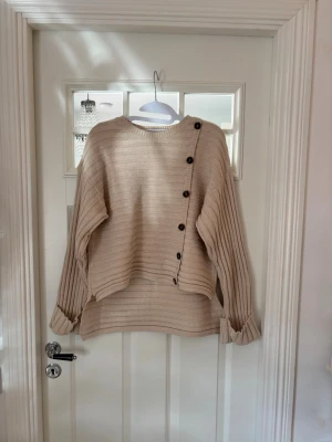 Beige stickad tröja med knappar - Säljer en beige stickad tröja med rund hals och dekorativa svarta knappar längs ena axeln och sidan. Tröjan har ribbade detaljer, långa ärmar och en avslappnad passform. Perfekt för dig som gillar en enkel men ändå unik stil.
