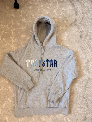 Grå hoodie från Trapstar med tryck - Snygg grå hoodie från Trapstar med broderat Trapstar-tryck i vitt och blått på bröstet. Klassisk huva med dragsko och ribbade muddar vid ärmar och nederkant. Tillverkad i mjuk bomullsblandning för skön känsla.