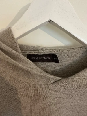 Kashmir från Oscar Jacobson - Säljer en stilren beige hoodie från Oscar Jacobson i mjukt stickat material. Hoodien har en klassisk huva och raka ärmar, perfekt för en avslappnad och snygg look. Passar dig som gillar minimalistisk design och hög kvalitet.
