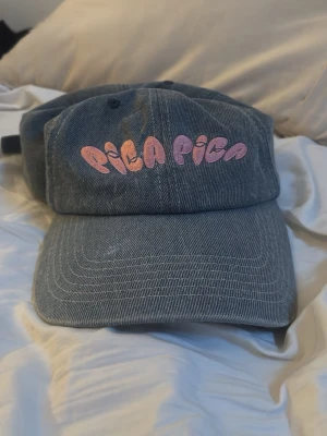 Grå keps från Pica Pica - Säljer en grå keps från Pica Pica med broderad rosa text framtill. Sjukt cool till en sommar outfit eller på stranden, älskar att den är jeans material!!
