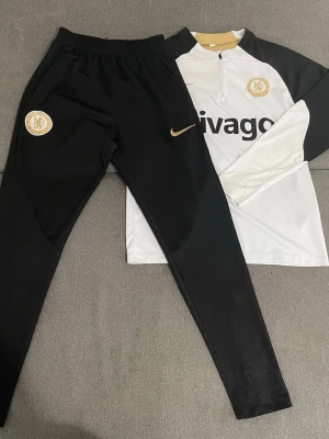 Chelsea träningsset från Nike - Chelsea FC träningsset från Nike med långärmad vit och svart tröja med halv dragkedja och byxor i svart. Tröjan har klubbmärke och sponsortryck, samt gulddetaljer. Tillverkad i lätt och funktionellt material, perfekt för fotbollsträning.