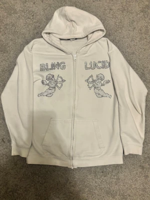 Vit hoodie från Bling Lucid med paljett tryck - Vit hoodie med dragkedja från Bling Lucid. På bröstet finns paljett tryck med texten 'BLING' och 'LUCID' samt två änglar i svart. Hoodien har fickor framtill och är gjord i mjuk bomull. Perfekt för en avslappnad och trendig stil. Finns ett litet håll vid fickan(se på bild 3) och några paljetter har lossnat men ingen stor skada. Storlek Kids XL