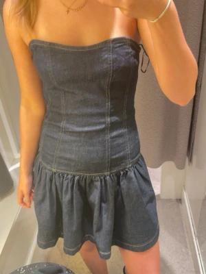 Mörkblå axelbandslös jeansklänning - Trendig mörkblå axelbandslös jeansklänning med markerad midja och utställd kjol. Klänningen har synliga sömmar som ger en cool look och är perfekt för dig som gillar denim. Passformen är normal och materialet är klassiskt jeans.