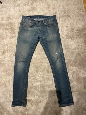 Blå slitna skinny/slim jeans - Säljer ett par blå dondup george jeans med slitningar och på ena benet. Jeansen har klassisk femficksdesign, låg midja och är tillverkade i denim med stretch. Perfekta för en avslappnad och trendig look.