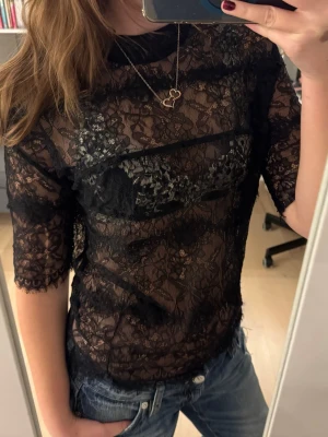 Svart spetsblus med halvärm - Snygg svart spetsblus med halvärm och hög hals. Blusen är helt genombruten i spetsmaterial och har fina spetsdetaljer vid ärmslut och nederkant. Perfekt att styla med jeans eller kjol för en trendig look.