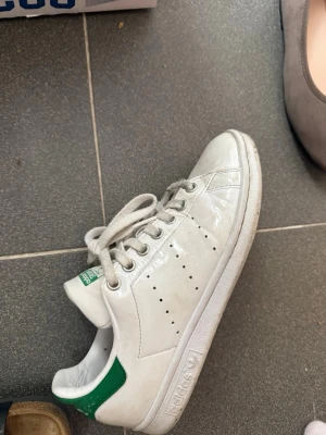 Adidas Stan Smith vita sneakers - Klassiska vita Adidas Stan Smith sneakers med gröna detaljer på hälen. Skorna har lågt skaft, perforerade ränder på sidorna och är gjorda i läder med gummisula. Perfekta för en clean och tidlös stil.