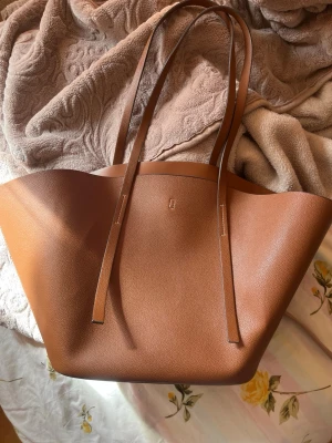 Brun tote bag i skinnimitation - Stilren brun handväska i tote-modell med långa axelband och minimalistisk design. Väskan är rymlig och har en mjuk struktur, perfekt för att bära allt du behöver. Tillverkad i skinnimitation med slät yta och diskret präglad logga framtill.