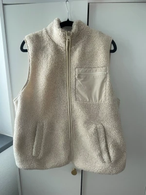 Krämvit teddyväst från Lindex - Mysig krämvit teddyväst från Lindex med dragkedja framtill, två sidofickor och en bröstficka med dragkedja i kontrasterande material. Hög krage och fluffig, fårullsinspirerad yta som ger en trendig look. Perfekt för lager-på-lager-stil under kyliga dagar. Knappt använd. Storlek L men liten i storlek
