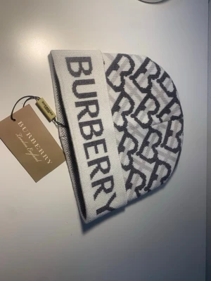 Bb mössa - Snygg vit mössa från Burberry med grått mönster och stor logga framtill. Tillverkad i mjukt stickat material som känns lyxigt och trendigt. Perfekt accessoar för dig som vill ha en exklusiv och modern look.