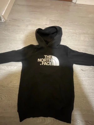 Svart hoodie från The North Face - Säljer en svart hoodie från The North Face i juniorstorlek XL. Hoodien har stor vit logga på bröstet och klassisk huva. Tillverkad i mjukt material, perfekt för chill dagar eller när du vill ha en clean streetwear-look.