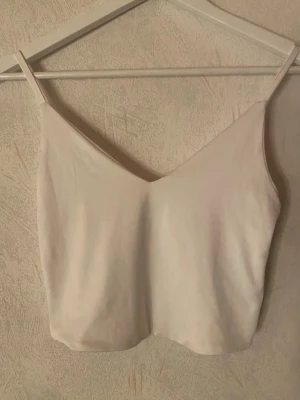 Vitt linne - Säljer en enkel och stilren vit linnetopp med smala axelband och v-ringad hals. Toppen har justerbara band och är perfekt för varma dagar eller som layering-plagg. Passar dig som gillar minimalistisk stil.
