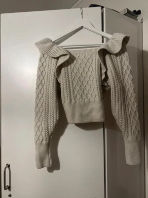 Beige stickad tröja med volang - Trendig beige stickad tröja med volangdetaljer vid axlarna och ett snyggt kabelstickat mönster. Tröjan har bred ribbad mudd i midjan och vid ärmsluten samt långa ärmar. 