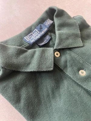 Grön långärmad piké från Polo Ralph Lauren - Mörkgrön långärmad pikétröja från Polo Ralph Lauren med klassisk krage och knappar framtill. Tröjan har en liten röd broderad logga på bröstet och är tillverkad i bomull för en bekväm känsla. Perfekt för en avslappnad men stilren look.