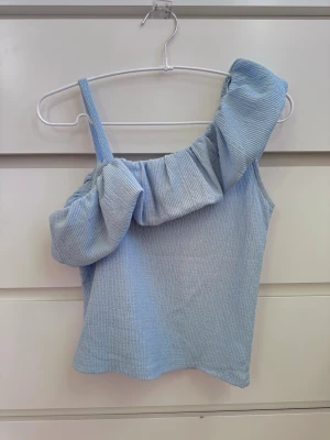 Ljusblå ribbad oneshoulder topp - Trendig ljusblå topp med ribbad struktur och oneshoulder-design. Toppen har en bred volang längs halsringningen och ett smalt axelband på ena sidan. Perfekt för dig som vill sticka ut med en unik och söt stil 😚🩵