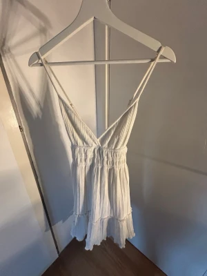 Vit kort sommarklänning med volang - Söt vit kort klänning med smala axelband och v-ringning. Klänningen har resår i midjan och volangdetaljer längst ner. Perfekt för varma dagar och har en luftig, avslappnad passform. Har en fläck!!!