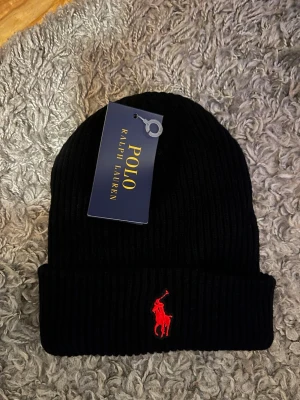 Svart ribbad mössa från Polo Ralph Lauren - Snygg svart ribbstickad mössa från Polo Ralph Lauren med klassisk röd broderad logga framtill. Mössan har uppvikt kant och är tillverkad i mjukt material som håller dig varm. Perfekt accessoar för kalla dagar och enkel att matcha med olika outfits.
