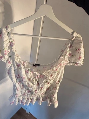 Blommig smockad topp från Gina Tricot - Supersöt vit topp från Gina Tricot med rosa blommigt mönster, smockad midja och volangkant. Toppen har korta puffärmar, söt rosettdetalj framtill och en croppad passform. Perfekt för sommarens alla tillfällen!