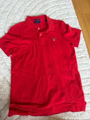 Röd pikétröja från Gant - Klassisk röd pikétröja från Gant med liten logga broderad på bröstet. Tröjan har krage, korta ärmar och knappar vid halsen. Tillverkad i mjuk bomull som känns skön mot huden. Perfekt till jeans eller shorts för en avslappnad look. Man får även med en gratis vara på sista bilden🤯🤯🤯