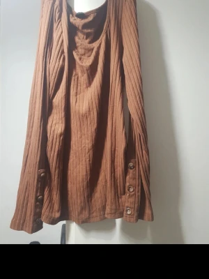 Brun ribbad långärmad topp - Snygg brun ribbad topp med långa ärmar och dekorativa knappar vid ärmsluten. Toppen har en lösare passform och en mjuk, något draperad halsringning. Perfekt för dig som gillar en avslappnad men trendig stil. Storlek:L