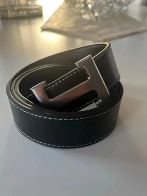 Svart skärp med H-spänne i skinn 115cm - Snyggt svart bälte i skinn med ett stort, silverfärgat H-format spänne. Bältet har vita kontrastsömmar längs kanterna som ger en clean och modern look. Perfekt accessoar för att lyfta vilken outfit som helst.