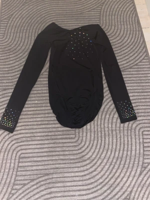 Svart body med färgglada strasstenar - Snygg svart body med långa ärmar och färgglada strasstenar på bröstet och vid ärmsluten. Materialet är stretchigt och följsamt, perfekt för en trendig look. Ingen synlig logga eller märke. Djupare ringning både fram och bak.