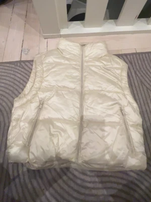 Krämvit dunväst från Softy, S - Stilren krämvit dunväst från Softy med hög krage och två dragkedjeförsedda fickor. Västen har en glansig finish och stängs med dragkedja framtill. Perfekt för lager-på-lager och ger en clean look till din outfit.
