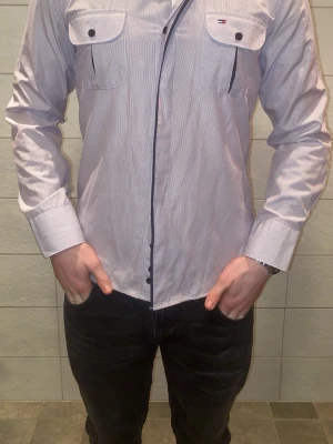skjorta från Tommy Hilfiger - Snygg rosa skjorta från Tommy Hilfiger med två bröstfickor och knappar framtill. Skjortan har lång ärm och diskreta ränder i tyget samt rosa detaljer vid fickorna. Perfekt för dig som gillar stilrena plagg med klassisk touch.