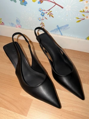 Svarta slingback pumps från NA-KD - Stilrena svarta pumps från NA-KD med spetsig tå och låg klack. Skorna har öppen häl med smal rem runt ankeln och är tillverkade i slätt skinn. Perfekta för dig som gillar en klassisk och elegant look. Aldrig använda💕