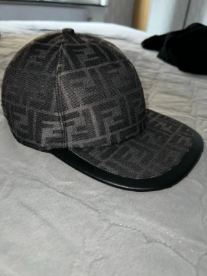 Svart keps med Fendi-logga - Säljer en svart keps från Fendi med det klassiska FF-mönstret över hela kepsen. Kepsen har böjd skärm i svart och justerbart band bak med Fendi-logga i guld. Materialet är textil med detaljer i konstläder på skärmen. Perfekt för dig som gillar exklusiva accessoarer. PRIS EJ HUGGET I STEN