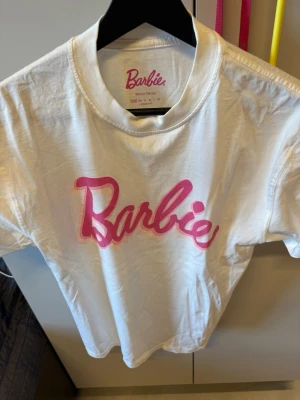 Vit Barbie t-shirt med rosa tryck - Säljer en vit t-shirt från Barbie med stort rosa Barbie-tryck på bröstet. Klassisk rund halsringning och korta ärmar. Fint till sommaren. Står xxs men passar xs och s