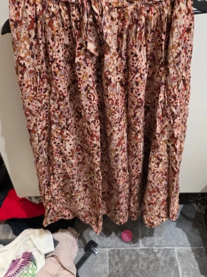 Mönstrad långkjol med knytband - Säljer en flowy långkjol med abstrakt mönster i beige, brun, röd och rosa toner. Kjolen har ett knytband i midjan och är gjord i ett lätt, mjukt tyg som passar perfekt till varmare dagar.