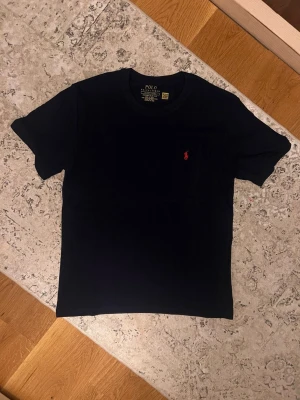 Svart t-shirt från Polo Ralph Lauren - Snygg svart t-shirt från Polo Ralph Lauren med klassisk röd broderad logga på bröstet. T-shirten har rund halsringning och korta ärmar. Perfekt basplagg med stilren design och skön passform.