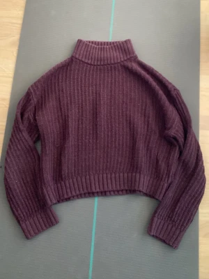 Vinröd ribbstickad polotröja - Mysig vinröd ribbstickad polotröja med croppad passform och långa ärmar. Tröjan har breda muddar vid ärmslut och nederkant samt en hög krage som ger extra värme. Perfekt för lager på lager och en trendig look.