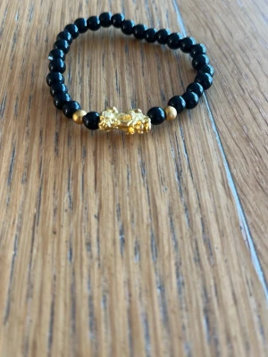 Svart pärlarmband med gulddetalj - Snyggt armband med svarta pärlor och små guldfärgade detaljer. I mitten sitter en dekorativ guldfärgad figur som ger armbandet en unik touch. Perfekt accessoar för dig som gillar stilrena men ändå iögonfallande smycken.