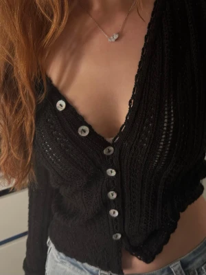 Svart virkad kofta med v-ringning - Trendig svart virkad kofta med djup v-ringning och genomskinlig struktur. Koftan har långa ärmar och vita knappar framtill som kontrast. Perfekt att styla med jeans eller kjol för en cool look.