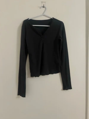 Grå v-ringad långärmad tröja  - En grå  långärmad tröja med v-ringning och lätt ribbad struktur. Toppen har en figurnära passform och vågig kant nertill samt vid ärmslut. Perfekt att styla med jeans eller kjol för en trendig look.