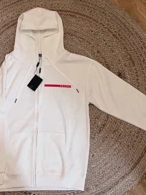 Vit hoodie med dragkedja från Prada - Snygg vit hoodie från Prada med dragkedja framtill och huva med dragsko. På bröstet finns en diskret rosa logga med texten PRADA. Hoodien har känguruficka och stilren design, perfekt för en trendig och avslappnad look.