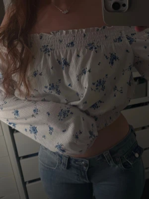 Offshoulder blommig topp i vitt och blått - Söt offshoulder topp med blå blommigt mönster på vit botten. Toppen har långa ärmar och smockdetaljer upptill och nedtill för en bekväm passform. Perfekt till jeans eller kjol för en trendig look.