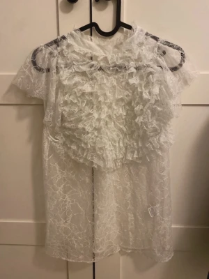 Vit transparent spetsblus med volanger - Säljer en vit, transparent blus i spets med korta ärmar och volangdetaljer framtill. Blusen har en luftig och romantisk look med ett unikt mönster i tyget. Perfekt för dig som gillar statement-plagg och vill sticka ut lite extra.