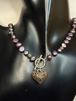 Romantiskt vintagehalsband från 90-talet med hjärthänge och äkta barocka sötvattenspärlor | Retro bohostil - Vintage halsband från svenska VÅGA i romantisk bohostil. Barocka sötvattenspärlor i djup brunlila ton kombineras med dekorativa metalldetaljer och ett genombrutet hjärthänge. Typisk 90-talsdesign med mjuk retrokänsla – fint både till vardag och fest.  Längd ca 42 cm + 5,5 cm förlängning. Hjärta ca 2,5 × 2,3 cm.  Fint skick, inga skador. Lätt patina på metallen ger en fin vintagekaraktär.  Jag prutar inte, men erbjuder bra rabatt vid köp av flera saker. Kika gärna på mina andra annonser också.