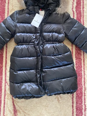 Svart dunjacka med pälshuva Moncler - Snygg svart dunjacka från Moncler med fluffig pälskant på huvan, använder inte eftersom den var för liten då jag fick den i present 💖 Jackan har en glansig finish, dragkedja framtill och markerad midja för en schysst siluett. Den är i storlek 14 år men kan möjligtvis passa tjejer som bär xs. 🫶🏽