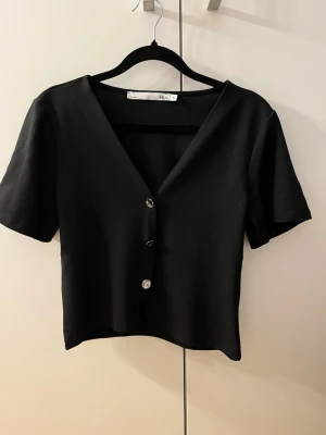 Svart kortärmad topp från Lager 157 - Stilren svart topp från Lager 157 med v-ringning och tre knappar framtill. Kortärmad modell i mjukt material som sitter snyggt och passar till många olika looks. Enkel och clean design som är lätt att matcha.