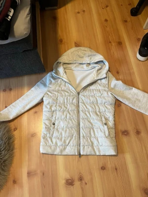 Ljusgrå quiltad hoodie-cardigan - Ljusgrå cardiganjacka med quiltad front och mjuka ärmar samt huva. Jackan har dragkedja framtill och två fickor med knappstängning. Materialet är en mix av vadderad polyester på framsidan och sweatshirt-tyg på ärmar och huva. Perfekt för lager-på-lager.