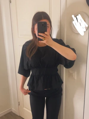 Svart topp med volang och markerad midja - Säljer en svart topp med bred resår i midjan och volang nedtill. Toppen har rund halsringning och halvlånga ärmar med lös passform. Perfekt att styla med jeans eller kjol för en trendig look. Använd en gång 