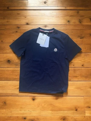 Mörkblå Moncler t-shirt - Snygg mörkblå t-shirt från Moncler med klassisk rund hals och korta ärmar. T-shirten har Monclers ikoniska logga broderad på bröstet och en diskret randig detalj i halsen. Tillverkad i mjuk bomull för en skön känsla.
